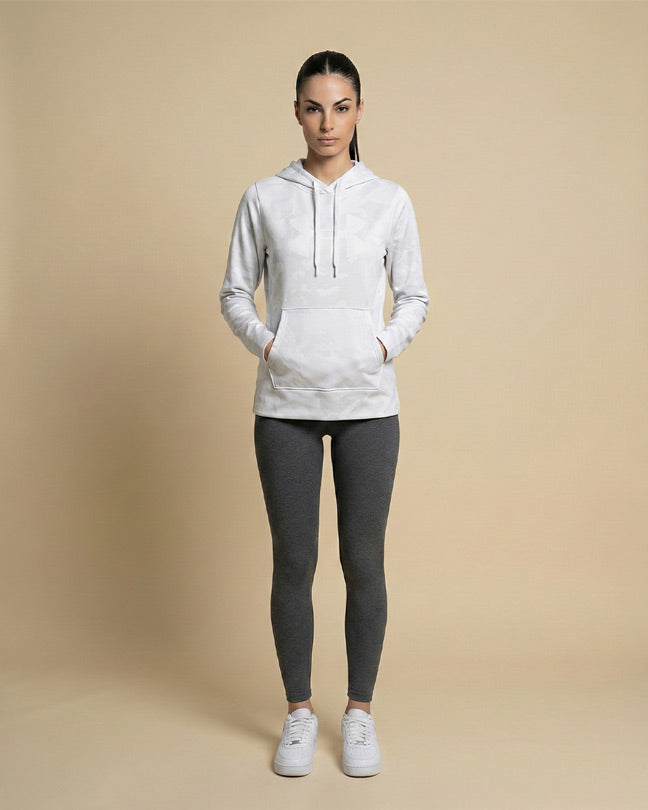 Woman wearing a light gray hoodie, dark leggings, and white sneakers standing against a beige background