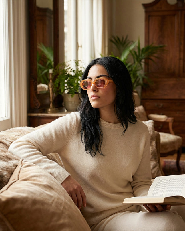 Woman in cream sweater wearing orange sunglasses, sitting on a sofa with a book in a cozy living room