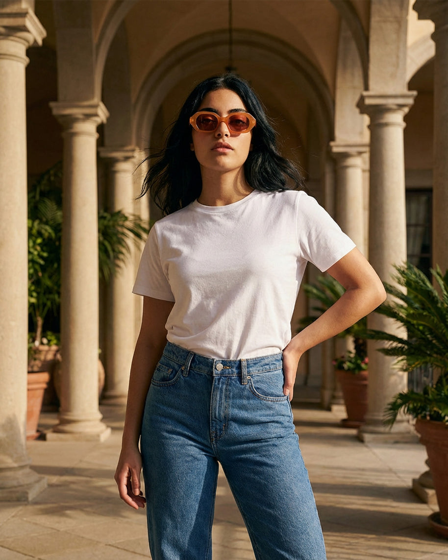 Woman in a white t-shirt and blue jeans wearing sunglasses, standing in a sunny courtyard with columns.
