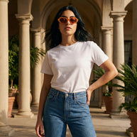 Woman in a white t-shirt and blue jeans wearing sunglasses, standing in a sunny courtyard with columns.