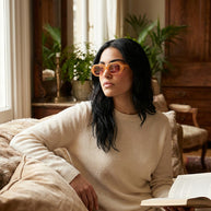 Woman in cream sweater wearing orange sunglasses, sitting on a sofa with a book in a cozy living room