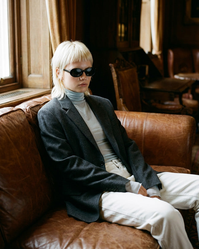 Person with blond hair and sunglasses wearing a gray blazer and turtleneck on brown sofa