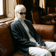 Person with blond hair and sunglasses wearing a gray blazer and turtleneck on brown sofa