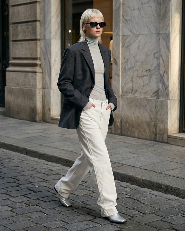 Woman in sunglasses, gray blazer, white jeans, and boots walking on city street