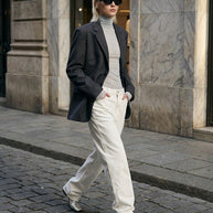 Woman in sunglasses, gray blazer, white jeans, and boots walking on city street