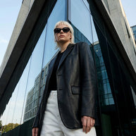 Woman in black leather blazer, white pants, and sunglasses standing by modern glass building