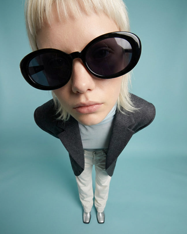 Fashionable woman in oversized round black sunglasses, gray blazer, and light pants on blue background