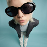 Fashionable woman in oversized round black sunglasses, gray blazer, and light pants on blue background