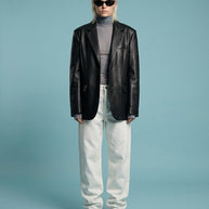 Person in a black leather blazer, gray turtleneck, white pants, and sunglasses on blue background