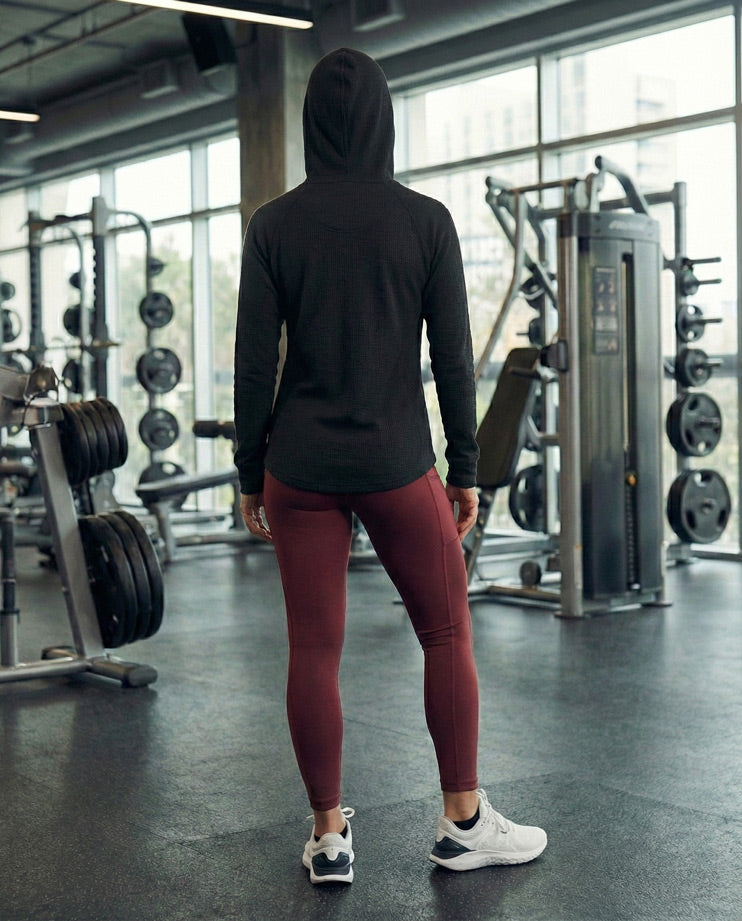 Person in black hoodie and maroon leggings standing in gym with workout equipment