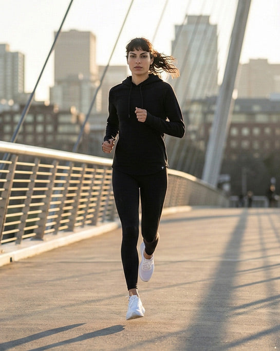 Woman jogging on bridge in black hoodie and leggings, city skyline in background.