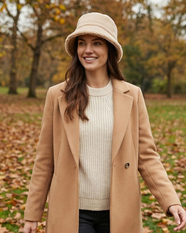 Woman in beige coat, knit sweater, and bucket hat standing outdoors in autumn park