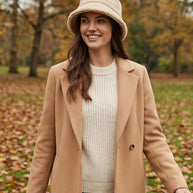 Woman in beige coat, knit sweater, and bucket hat standing outdoors in autumn park