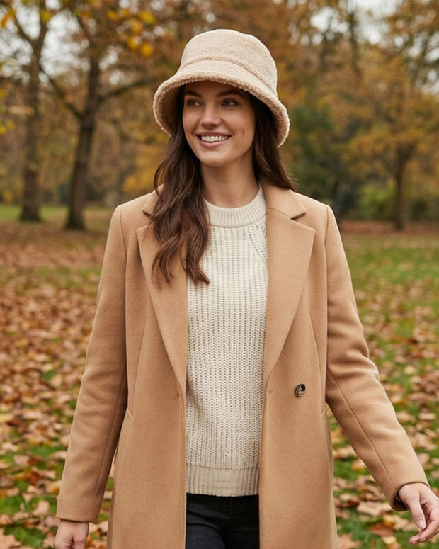 Woman in beige coat, knit sweater, and bucket hat standing outdoors in autumn park