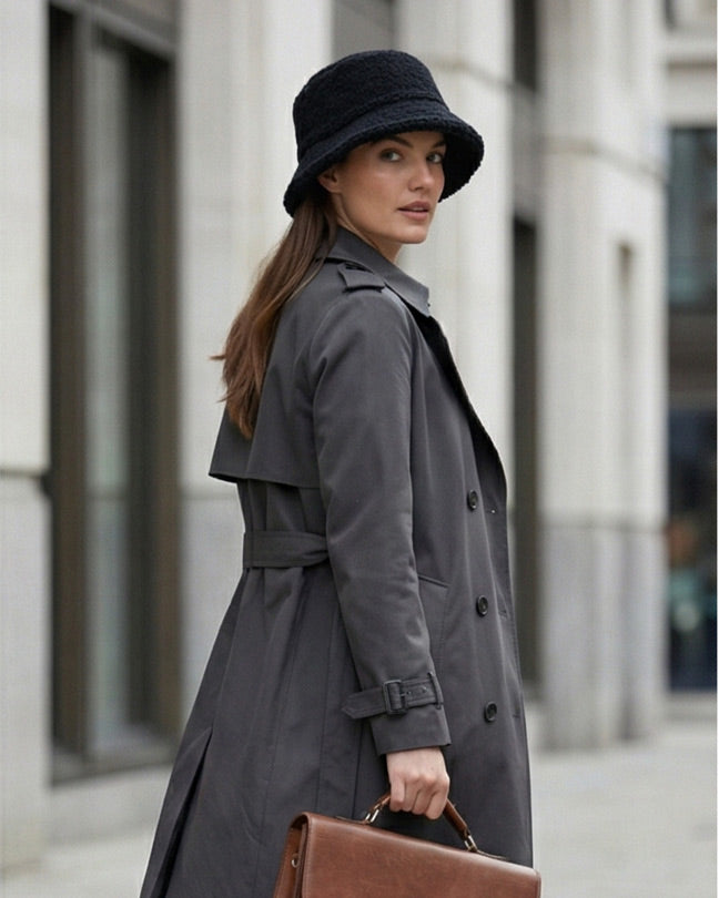Woman in gray trench coat and black bucket hat holding brown leather briefcase outdoors