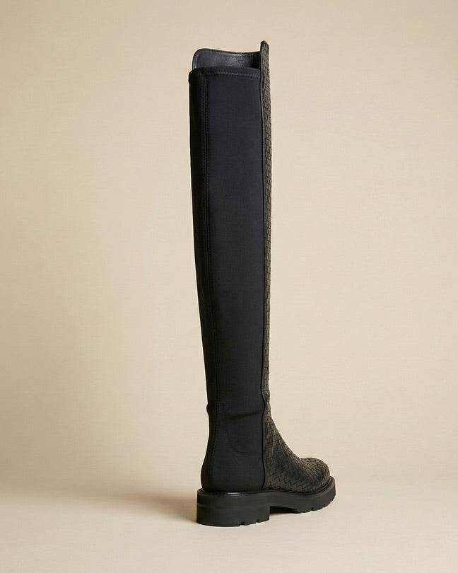 Black knee-high boot with textured pattern and thick sole on neutral background