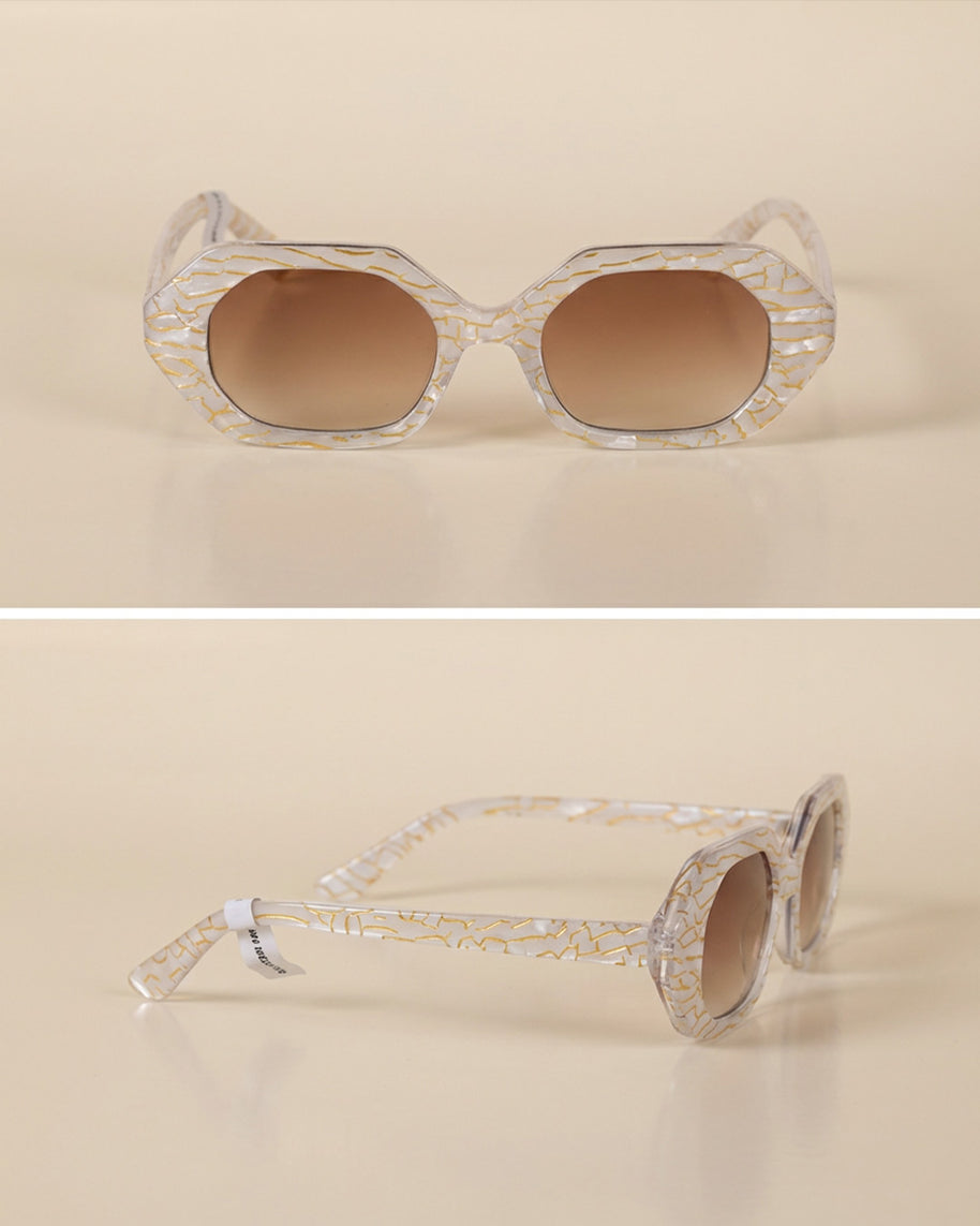 Geometric sunglasses with clear gold-patterned frames and brown gradient lenses on beige background