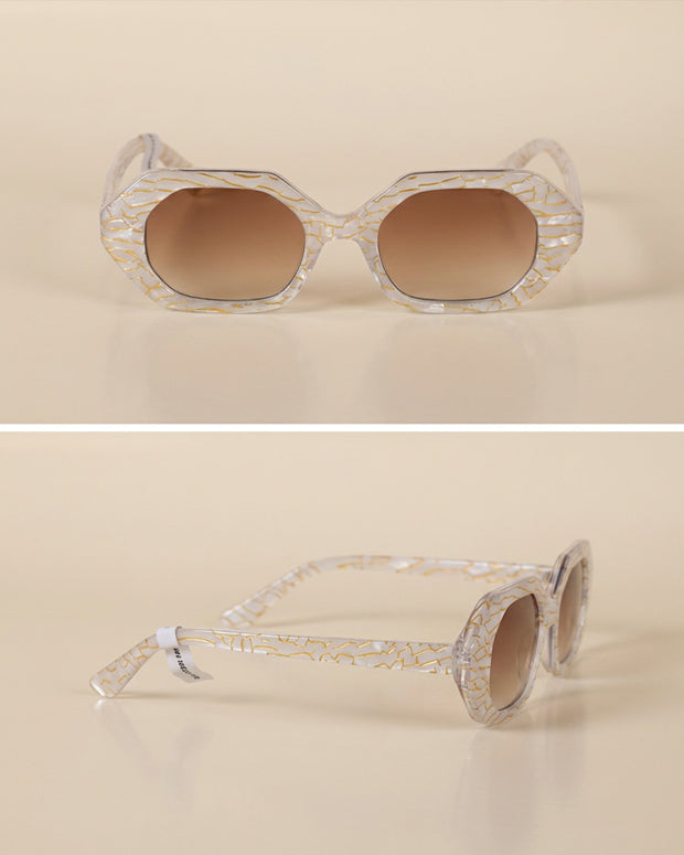 Geometric sunglasses with clear gold-patterned frames and brown gradient lenses on beige background
