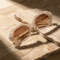 Beige geometric sunglasses with brown tinted lenses on textured stone surface in sunlight