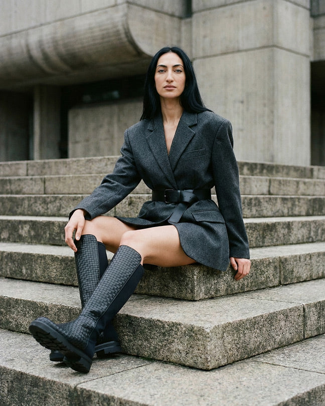 Woman in structured grey blazer and black knee-high boots sitting on stone steps outdoors