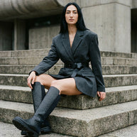 Woman in structured grey blazer and black knee-high boots sitting on stone steps outdoors