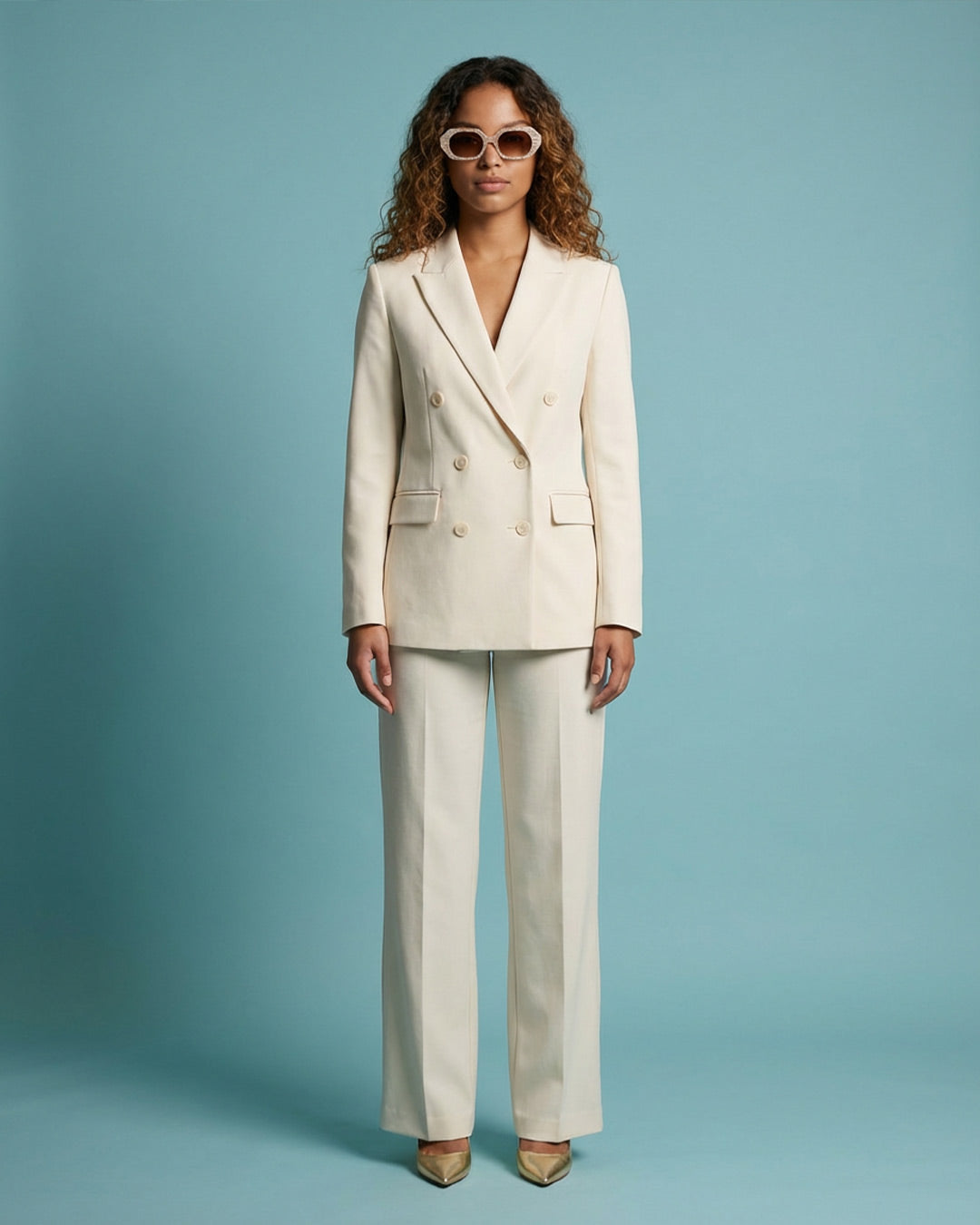 Woman in cream double-breasted pantsuit and sunglasses stands against a blue background