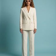 Woman in cream double-breasted pantsuit and sunglasses stands against a blue background